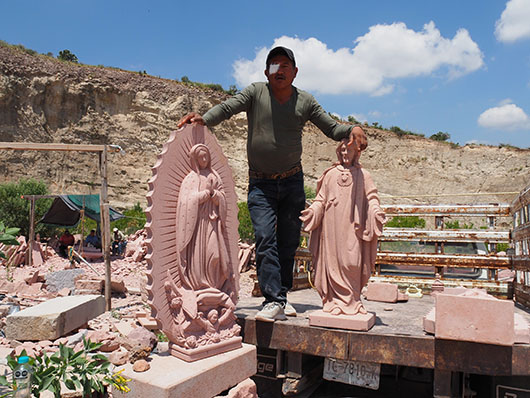 Man with an eye patch standing between two pink stone carvings of religious figures. Carver, Escallerias, San Luis Potosí. Photo by Sarah Lopez.