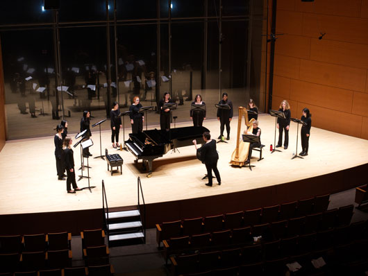 Camerata singers standing around a piano onstage