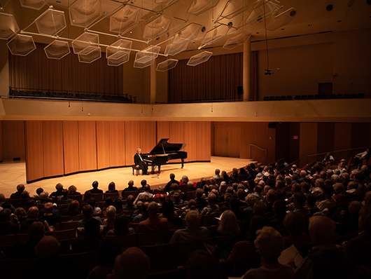 Jeffrey Siegel, seated at a grand piano, speaks to the audience in Pick-Staiger Concert Hall