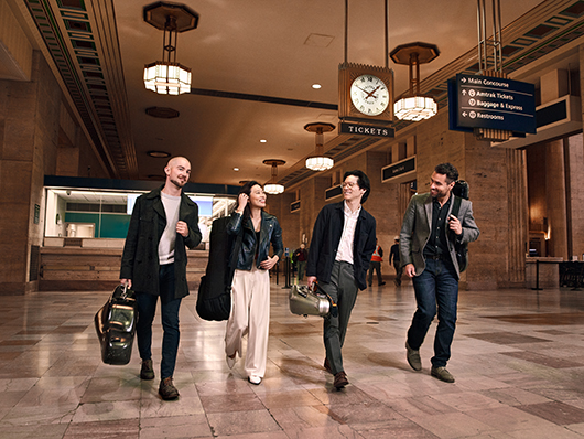 The members of the Dover Quartet walk through a train station, smiling and carrying their instrument cases