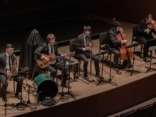 Musicians play seated onstage playing saxophone, guitar, woodwinds, cello, and violin.