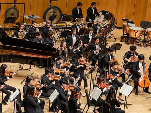 An orchestra of musicians play on stage, including strings, piano, woodwinds, and percussion.