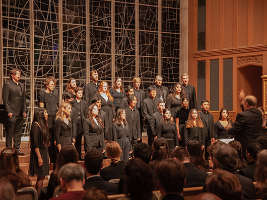 A choir dressed in black sings on a church stage in front of stained glass windows.