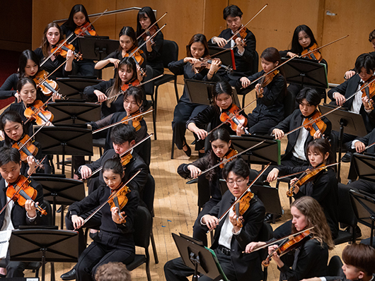 The violin section of an orchestra plays on a wooden stage.