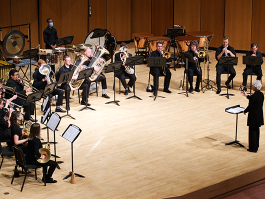 A semicircle of brass players perform as a conductor leads from the front.