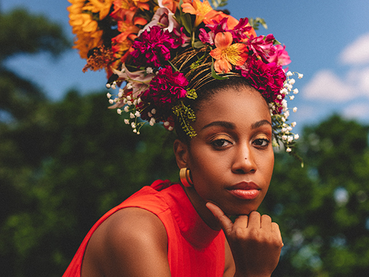 Jazzmeia looks contemplatively into the camera, with an elaborate and colorful flower arrangement on her head.