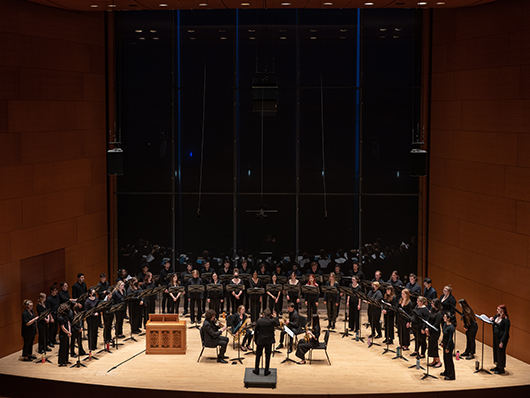 A conductor leads a large choir in a semicircle, accompanied by a small group of brass musicians.