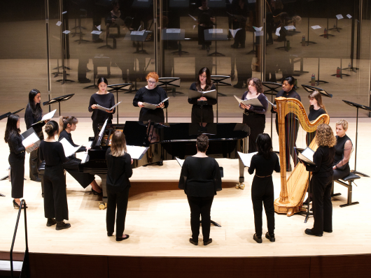 A circle of singers perform around a pianist.