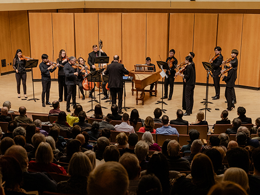 Musicians stand onstage performing behind a conductor, including violins, a double bass, a cello, and a harpsichord.