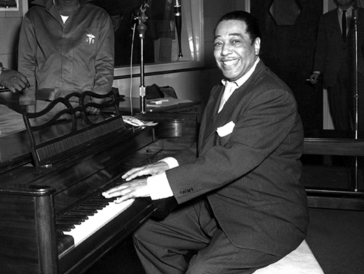 Duke Ellington sits at a piano, grinning broadly.