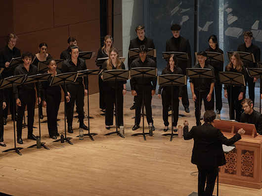 Two rows of singers perform on a wooden stage, led by a conductor and a pianist.