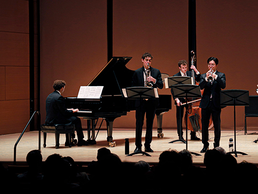 Four musicians play jazz on a stage, playing piano, saxophone, trumpet, and double bass.