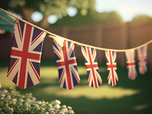 A line of Union Jack flags hangs over a sunny, grassy yard with a wooden fence.