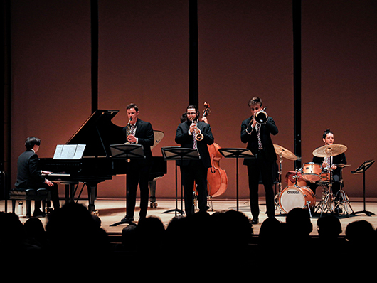 Four musicians play jazz onstage, including upright bass, trombone, drums, piano, and saxophone.