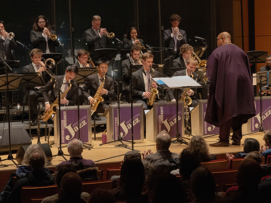 Rows of brass players play onstage as a conductor in a purple robe conducts.