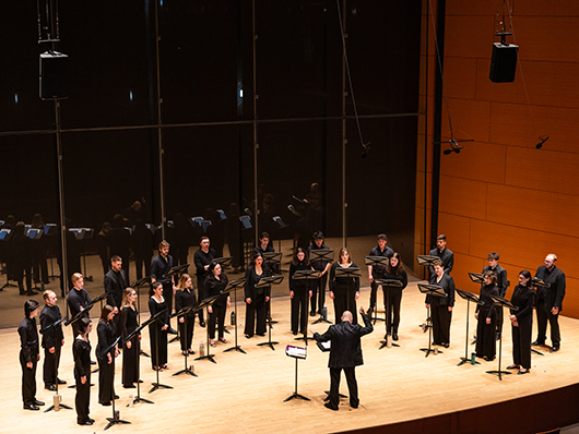 Two rows of singers stand in a semicircle, led by a conductor on a tall stage with floor to ceiling windows behind them.