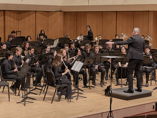 A band of woodwinds, brass, and percussionists play as a conductor leads.