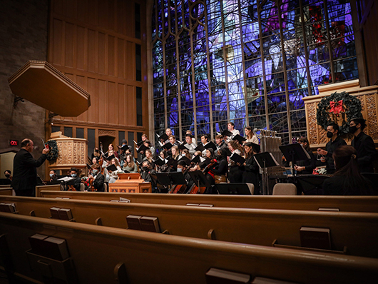 A choir sings on a church stage, led by a conductor and in front of a large blue stained glass window.