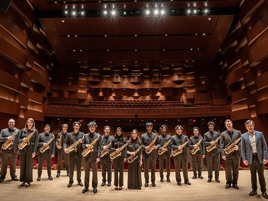 A large saxophone ensemble poses onstage with their saxophones.