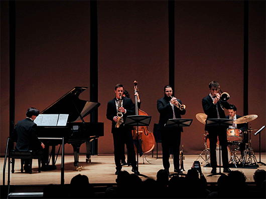 Three jazz musicians play onstage: a saxophonist, a trombonist, and a trumpeter. An upright bassist, drummer, and pianist play in the background.