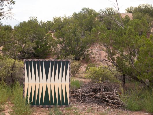 A painting with a symmetrical, geometric design of beige and dark green triangles is displayed in a naturally lush outdoor environment, surrounded by green plants, trees, and a pile of twigs.