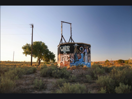 Old water tank on scrubland with text “Water is Life” and water-themed mural with three Diné people