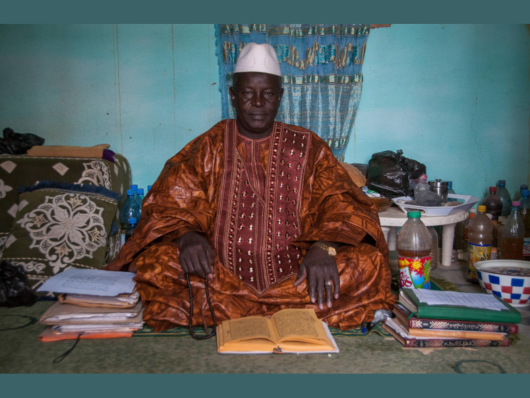 Dark-skinned man in brown embroidered robe sits cross-legged in crowded interior with manuscripts