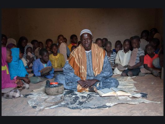 Dark-skinned man sits cross-legged with manuscript as boys and girls with Qur’an boards sit behind