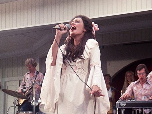 A woman with brown hair and light skin wearing a long white dress sings on stage