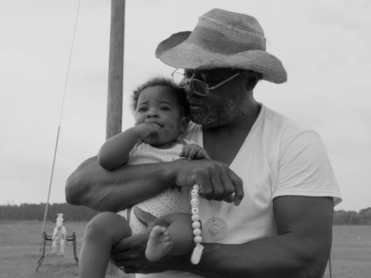 A man with dark skin tone and straw sun hat holding a baby outside