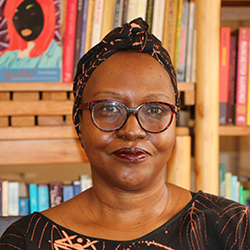 author Doreen Baingana wearing a brown patterned headscarf in front of a shelf filled with books