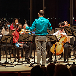 A conductor standing on stage and directing a group of string players.