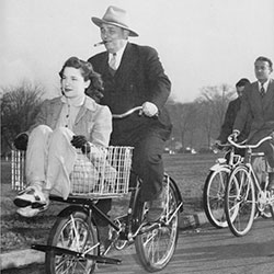 A black and white photo of a woman riding in a basket of a bicylce. The man on the bicycle is smoking a cigar and in a suit and hat. 