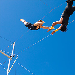 A trapeze artist flies in blue sky toward outstretched hands