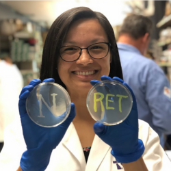 A lady holding up two petri dishes that say "N" and "RET"
