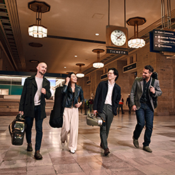 The members of the Dover Quartet walk through a train station, smiling and carrying their instrument cases