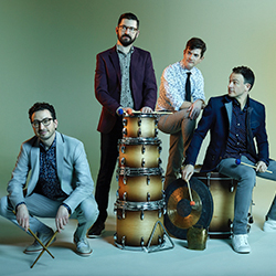 The members of Third Coast percussion stand in front of a green background with various percussion instruments