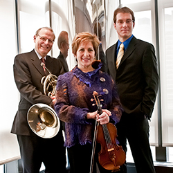 The members of Trio Valtorna stand holding their instruments and smiling in front of a window