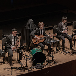 A saxophonist, guitarist, and woodwinds player perform seated onstage.