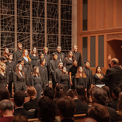 A choir dressed in black sings on a church stage in front of stained glass windows.