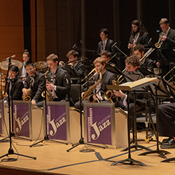 Jazz orchestra students perform seated onstage as a conductor in a purple robe conducts.