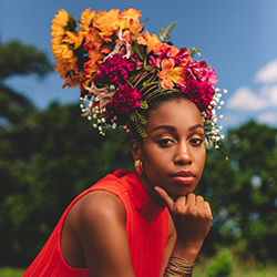 Jazzmeia looks contemplatively into the camera, with an elaborate and colorful flower arrangement on her head.