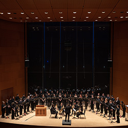 A conductor leads a large choir in a semicircle, accompanied by a small group of brass musicians.