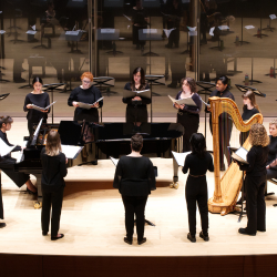 A circle of singers perform around a pianist.