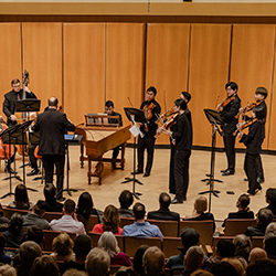 Musicians stand onstage performing behind a conductor, including violins, a double bass, a cello, and a harpsichord.