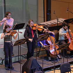 Musicians stand playing on a platform, including two cellists, two violinist, and a flautist.