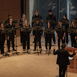 Two rows of singers perform on a wooden stage, led by a conductor and a pianist.