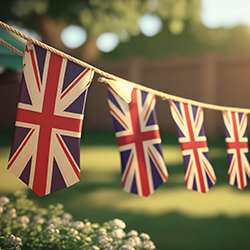 A line of Union Jack flags hangs over a sunny, grassy yard with a wooden fence.