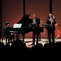 Four musicians play jazz onstage, including upright bass, trombone, drums, piano, and saxophone.