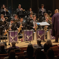 Rows of brass players play onstage as a conductor in a purple robe conducts.